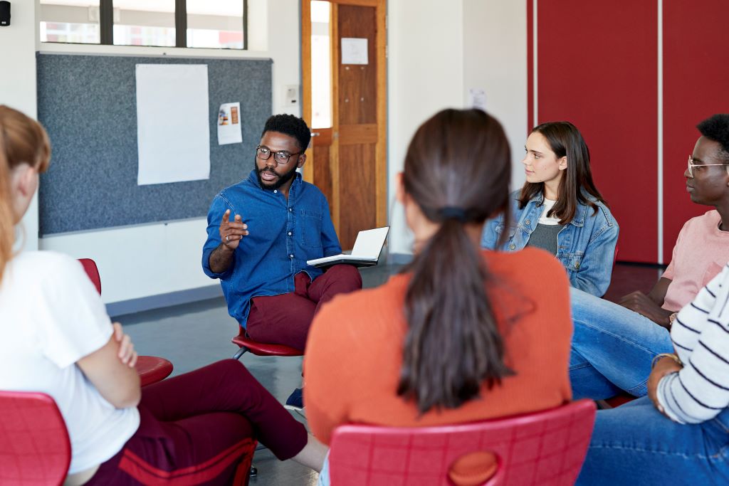 an instructor sitting in a class with a circle of students reading from a textbook.