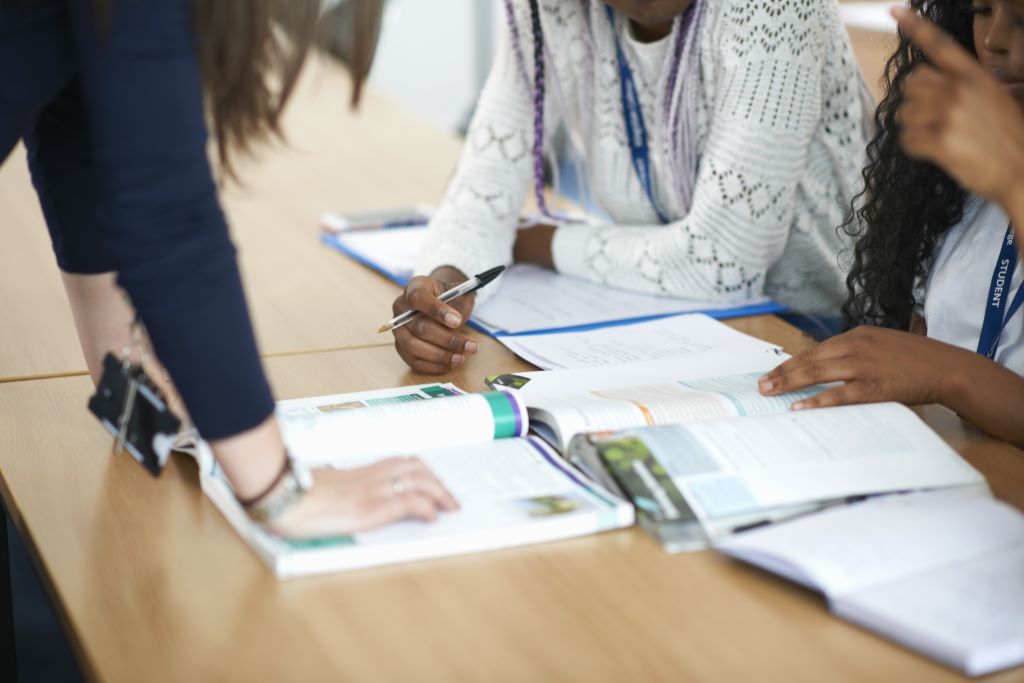 Three students in a library, working at a table, one is leaning against the table, they have several books open flat on the table.