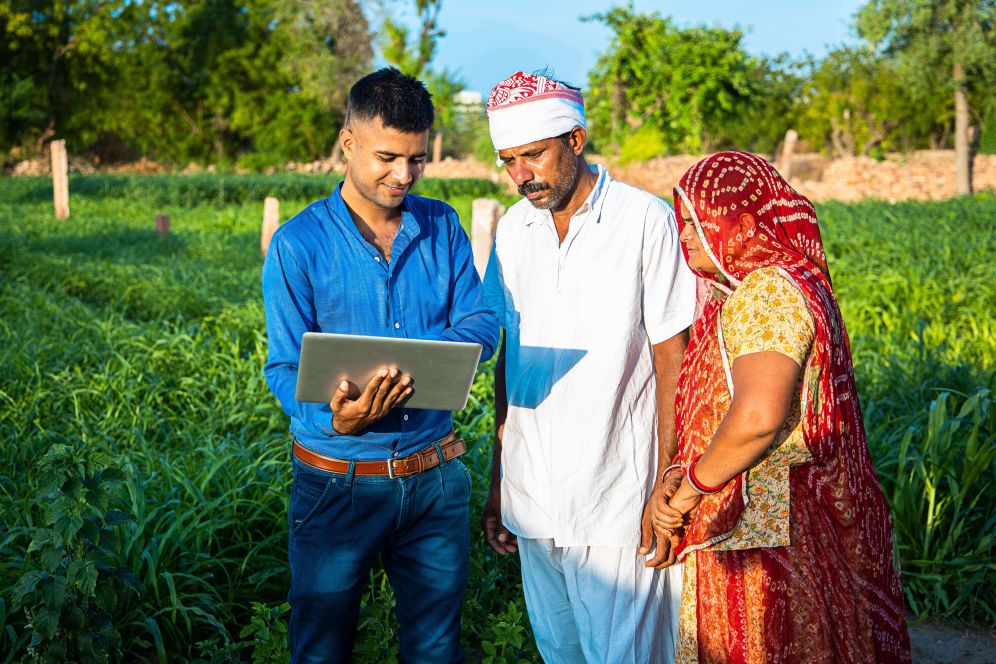 Young indian man with laptop showing something to village farmer family on internet standing in green field, agriculture land, teaching computer and use of technology.