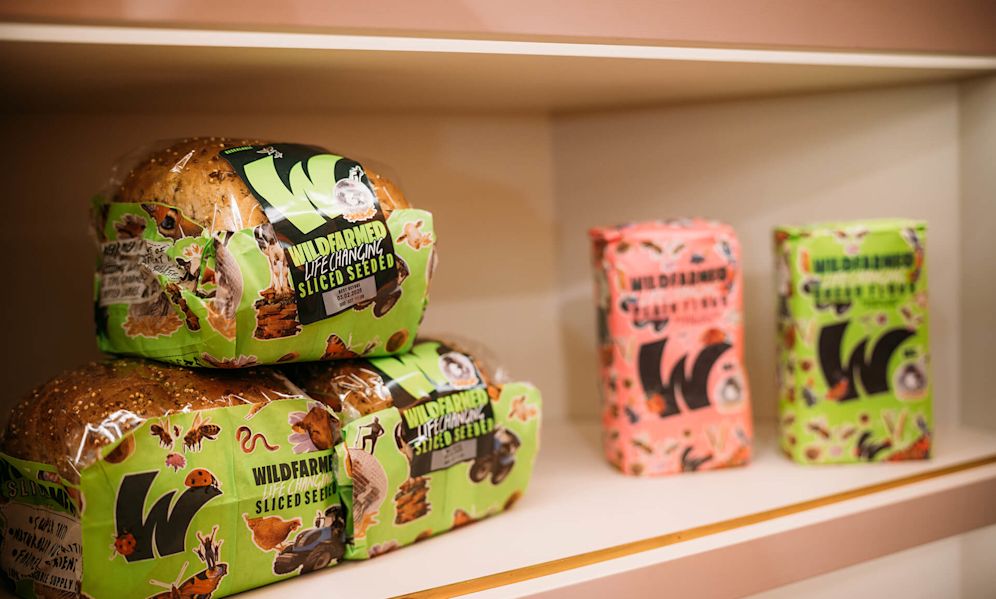 On a white shelf, several loaves of Wildfarmed brand bread are displayed. In the foreground, three loaves of 'Wildfarmed Sliced Seeded' bread are stacked, packaged in distinctive bright green wrapping with illustrations of various animals and plants. The seeded bread has visible whole grains on the crust. In the background, slightly out of focus, are two more Wildfarmed products - one in pink packaging and another in green packaging. All feature the prominent 'W' Wildfarmed logo. The products appear to be part of an environmentally-conscious bread line, displayed in what looks like a retail setting.