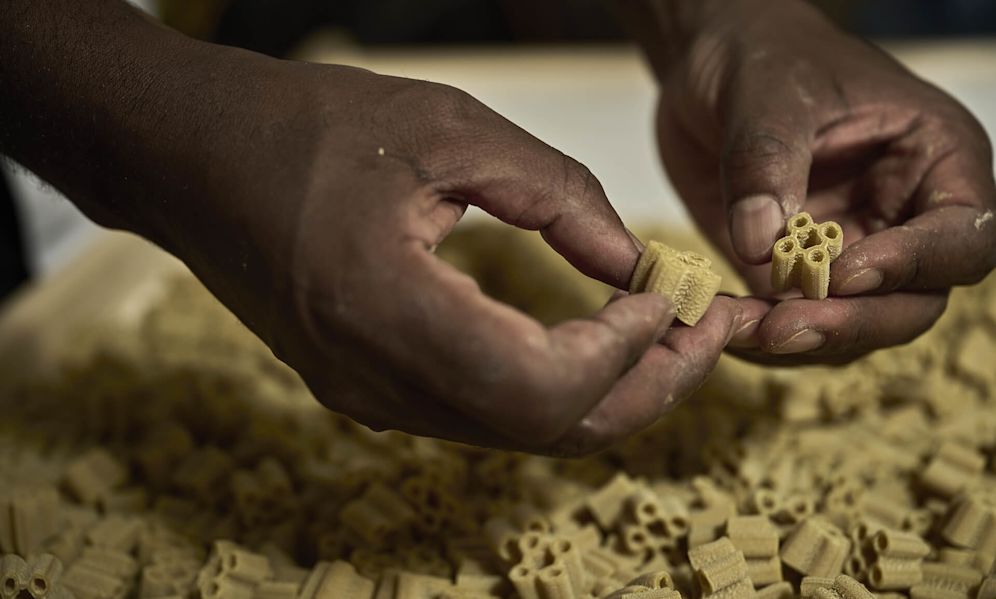 Close-up of hands examining small, yellow pasta pieces with unusual shapes and hollow centers. The hands are holding two different pasta shapes - one appears to be a curved tube and the other has multiple hollow sections.