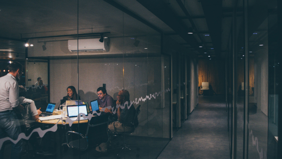 Office hallway with a meeting room on the left with glass windows. Inside the meeting room, people are sitting round a desk with laptops. 