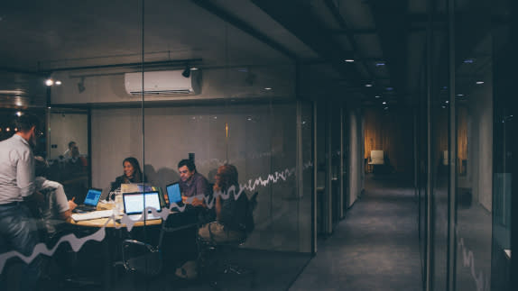 Office hallway with a meeting room on the left with glass windows. Inside the meeting room, people are sitting round a desk with laptops.