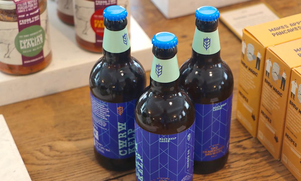 Three dark glass bottles of Hazy Kelp Lager beer with distinctive blue labels and mint green neck wraps. The bottles feature a geometric pattern and a wheat or barley symbol on the labels. They're arranged on a wooden table alongside food items including what appears to be sauce jars on the left and yellow packaging labeled 'Maker's Apple Pancakes' on the right.