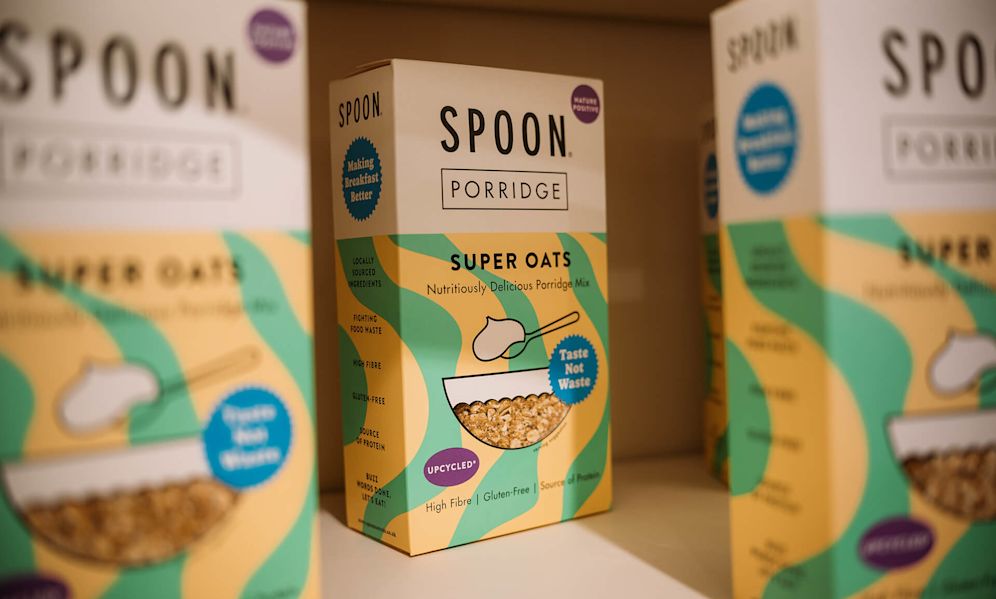 Here's alt text for this image:
"Boxes of SPOON brand porridge arranged on a shelf. The focal package in the center is labeled 'SPOON. PORRIDGE - SUPER OATS Nutritiously Delicious Porridge Mix' with a yellow and teal wavy pattern design. The packaging prominently features sustainability messaging including 'Upcycled,' 'Taste Not Waste,' 'Fighting Food Waste,' and 'Locally Sourced Ingredients.' An illustration shows a bowl of oats with a spoon. Additional product benefits listed include 'High Fibre,' 'Gluten-Free,' and 'Source of Protein.' Identical boxes appear slightly blurred in the background.
