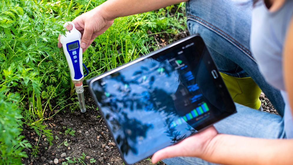 A close-up image of a PH meter tester inserted into soil. The tester's display shows the PH level, and the soil appears rich and dark. The image is set against a natural background, highlighting the importance of soil health and environmental monitoring.