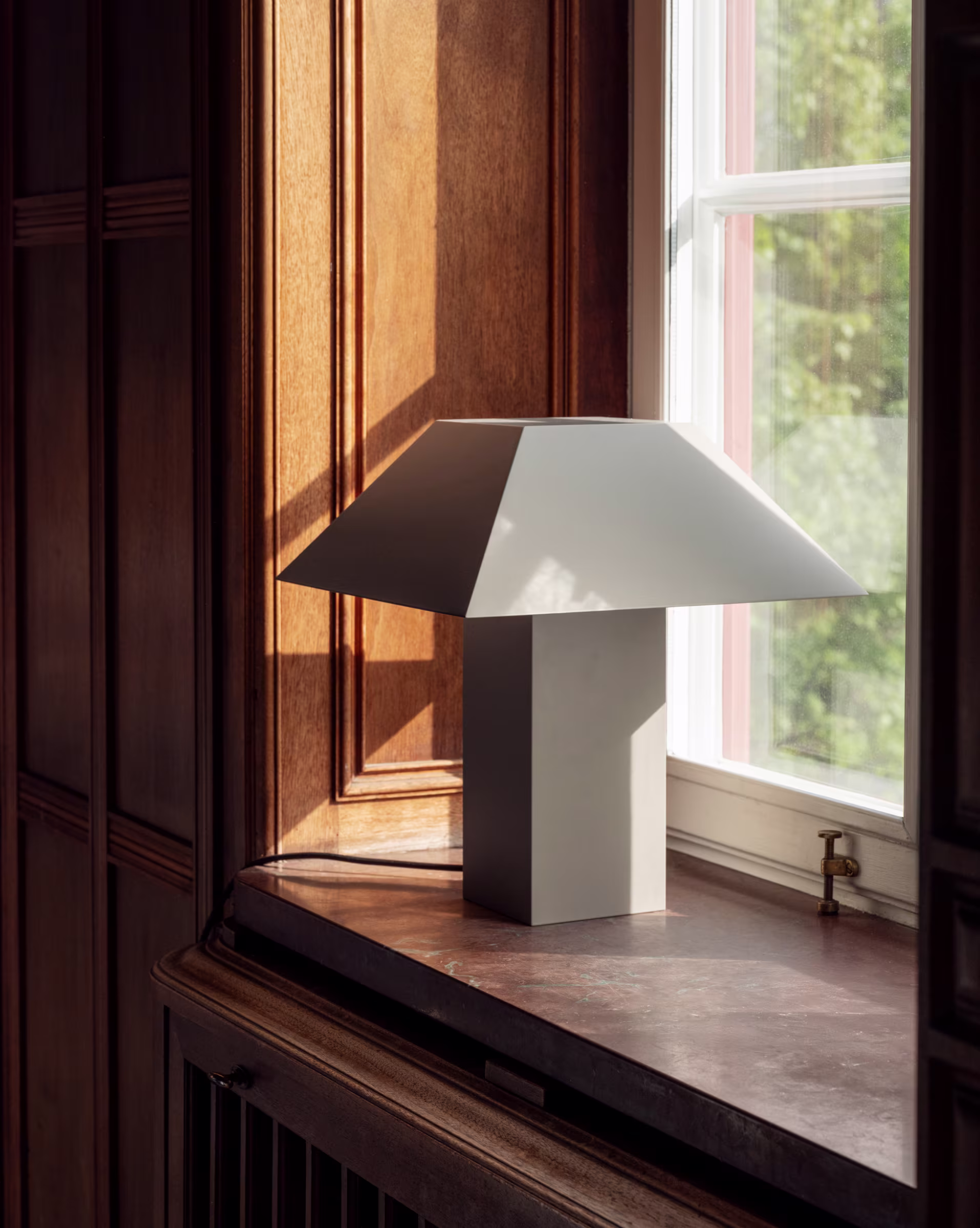 Lamp 53 av Axel Wannberg, en grå bordlampa, står på ett fönsterbräde i trä. Ett fönster med grönska utanför syns i bakgrunden.