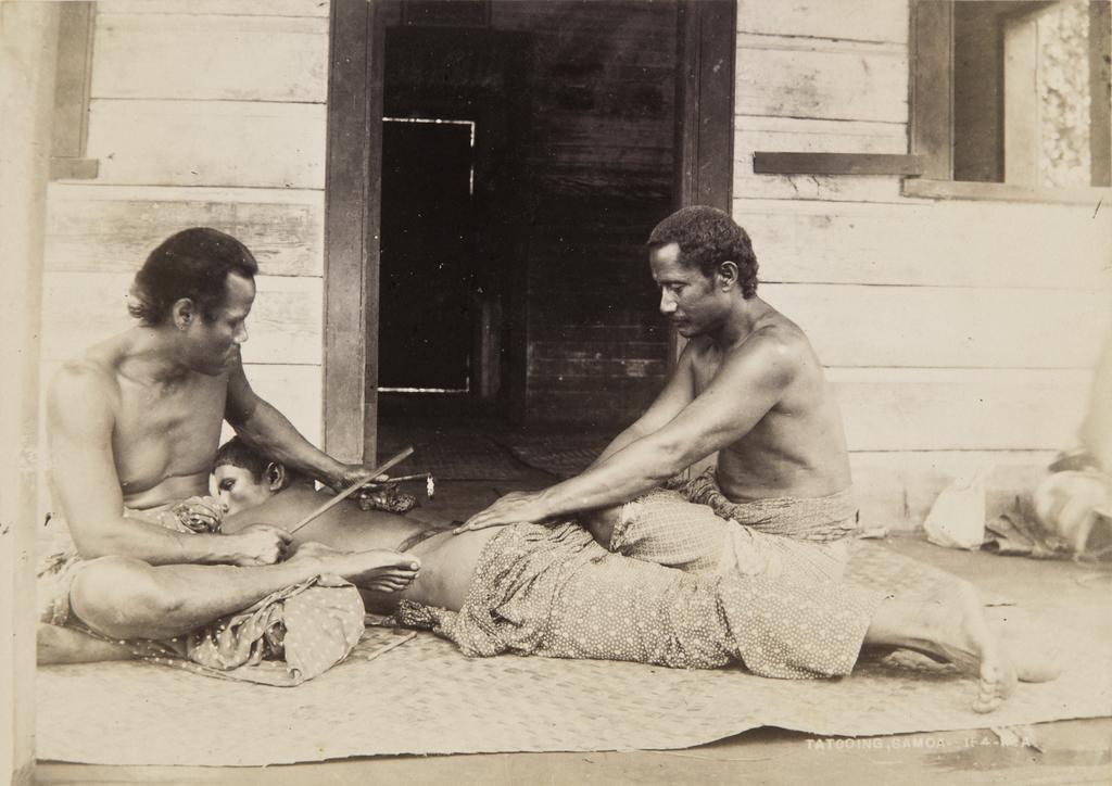 A young Samoan man receiving a tatau (tattoo) on the back by two others.