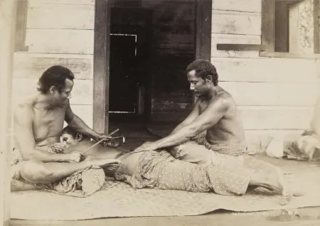 A young Samoan man receiving a tatau (tattoo) on the back by two others.