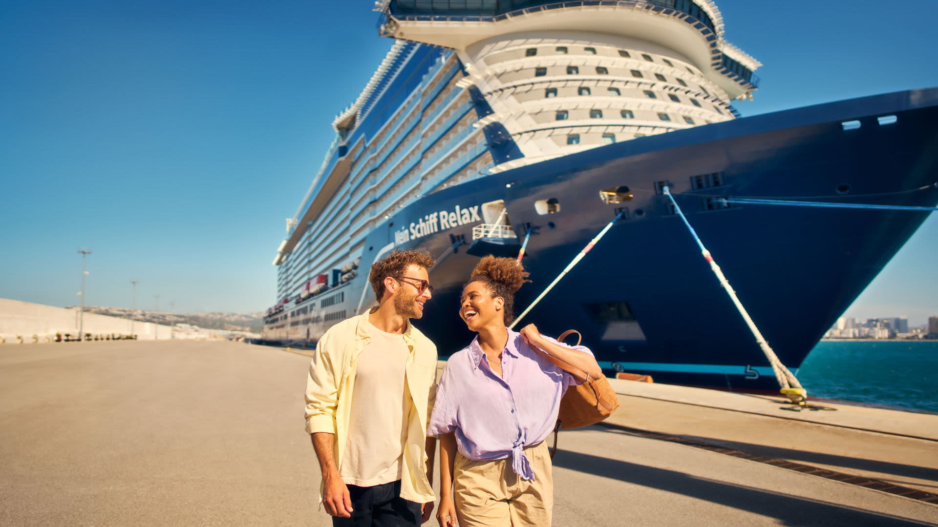Ein Pärchen spaziert am Anleger mit der Mein SchiffEndlich Zeit für eine neue Dimension des Wohlfühlens – z. B. an Bord der neuen Mein Schiff Relax  Relax im Hintergrund