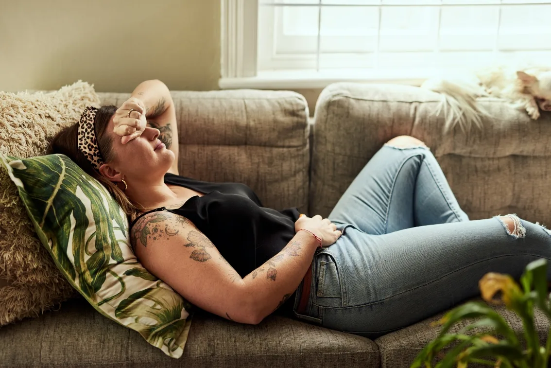 Woman with tattoos laying on couch in pain with her arm over her face