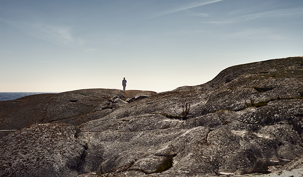 Utsikt over massive svaberg, blå himmel og sjø i bakgrunnen. En kvinne står og speider utover mot havet i det fjerne.