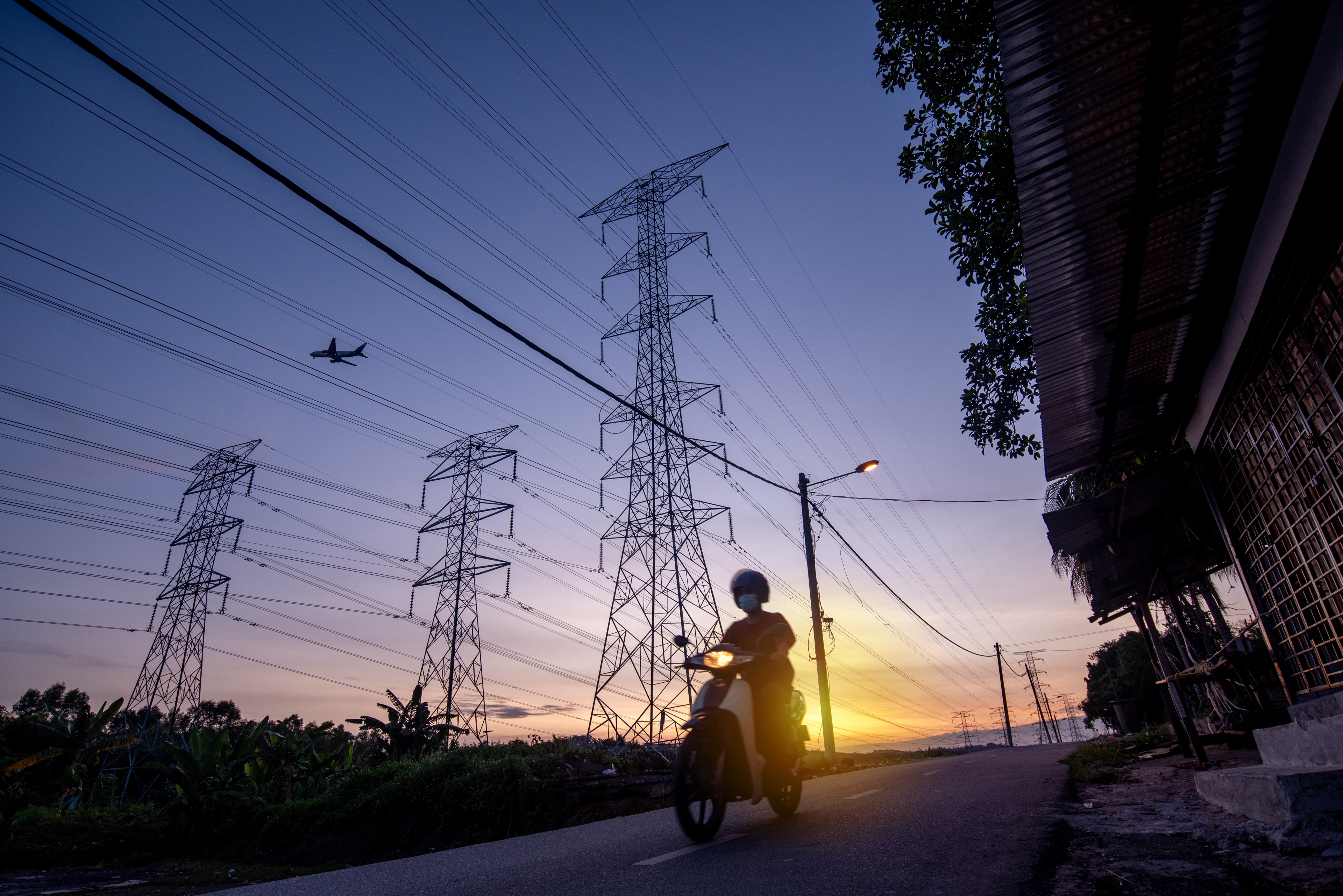 Electricity pylons at night with man on a motor scooter driving down the road to the backdrop of a sunset.