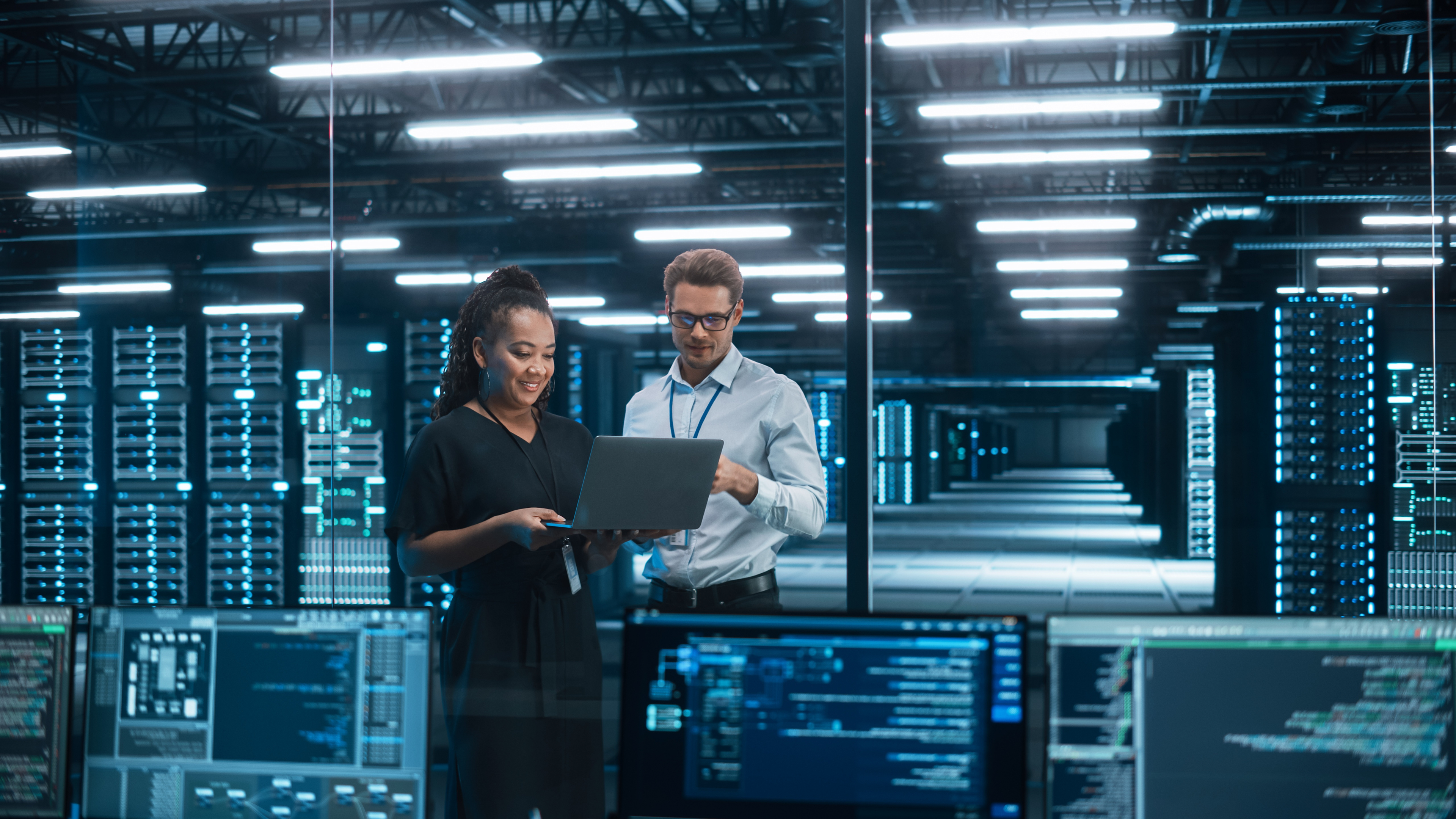 Business man and woman looking at computer in a large data centre, with racks of servers.