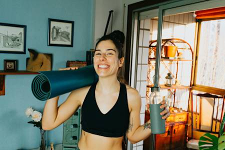 Smiling woman in black sports top holding a rolled yoga mat and water bottle in a bright, decorated home interior.