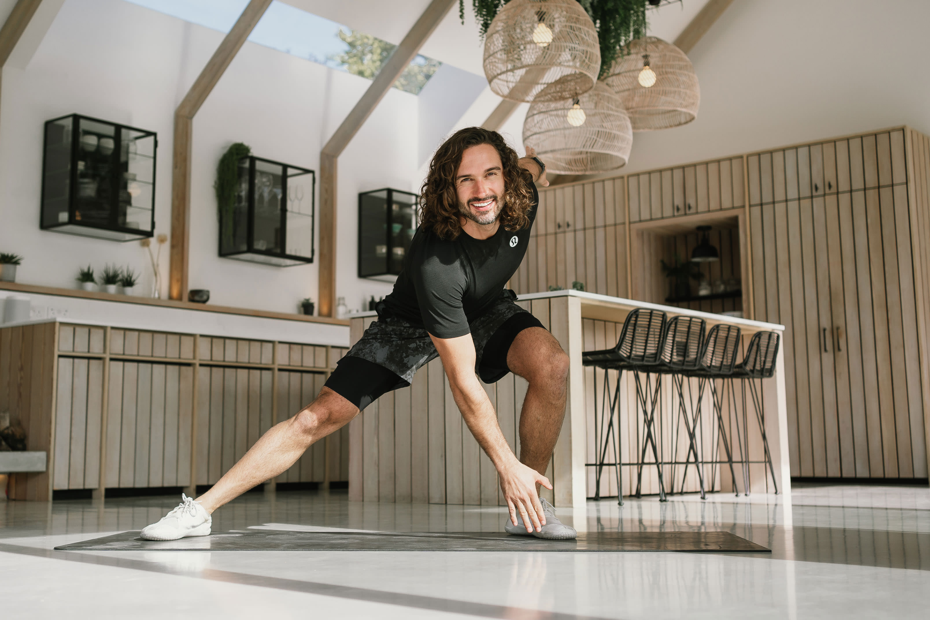 Joe Wicks wearing a black top reaches to the bottom of an exercise mat inside a kitchen.