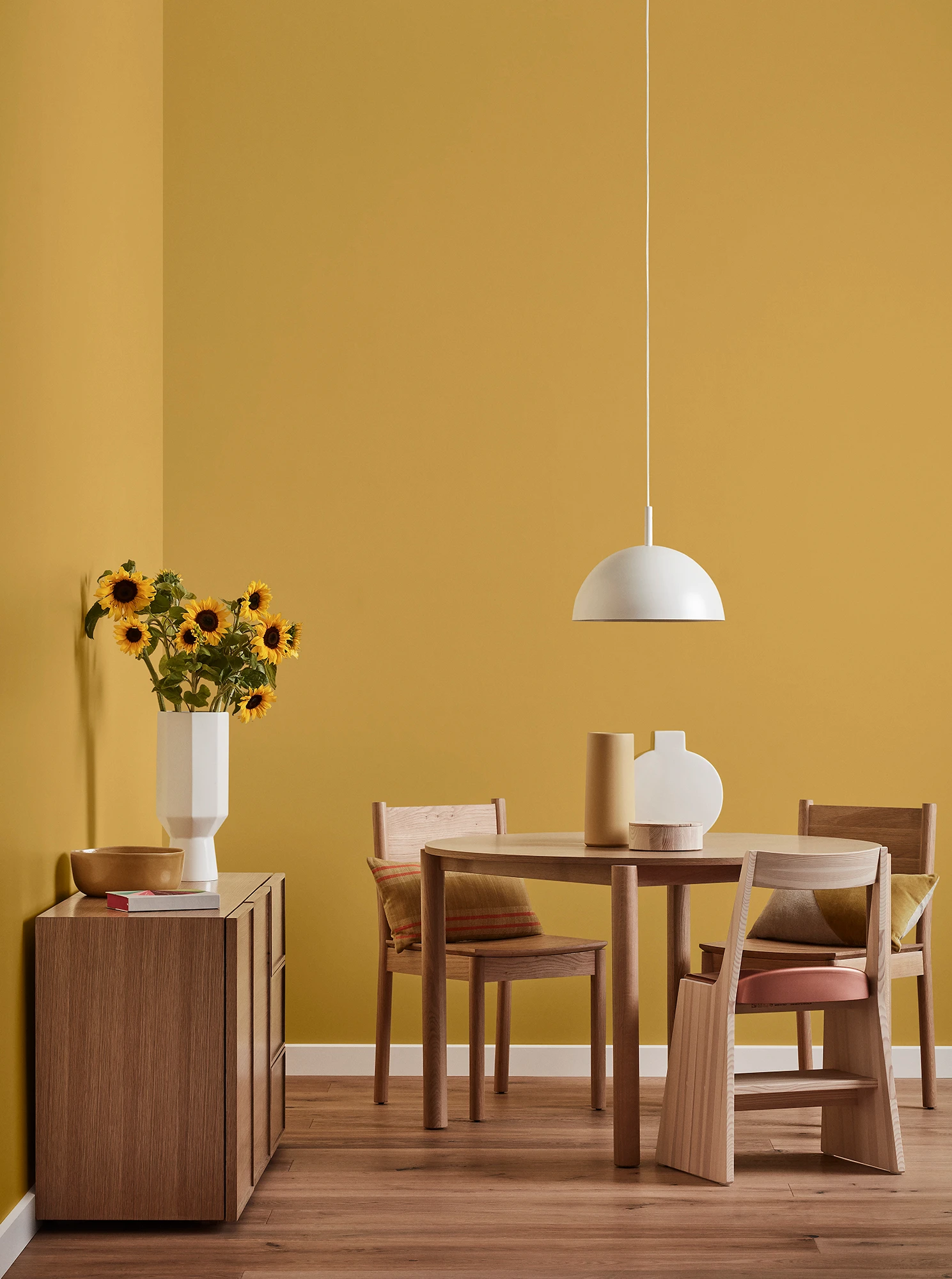 Round wooden table, three chairs and console with vase of sunflowers and white pendant light in mustard-yellow oom.