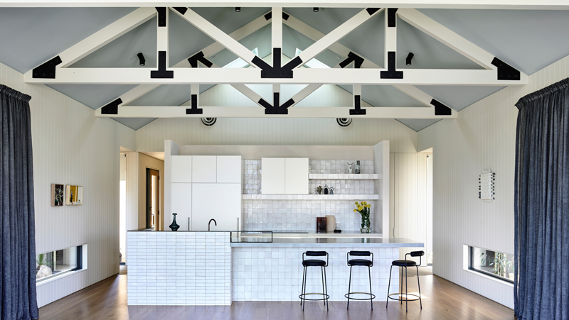 White tiled kitchen with island bench and three black stools. Light blue pitched ceiling. White exposed beams with black anchor points.
