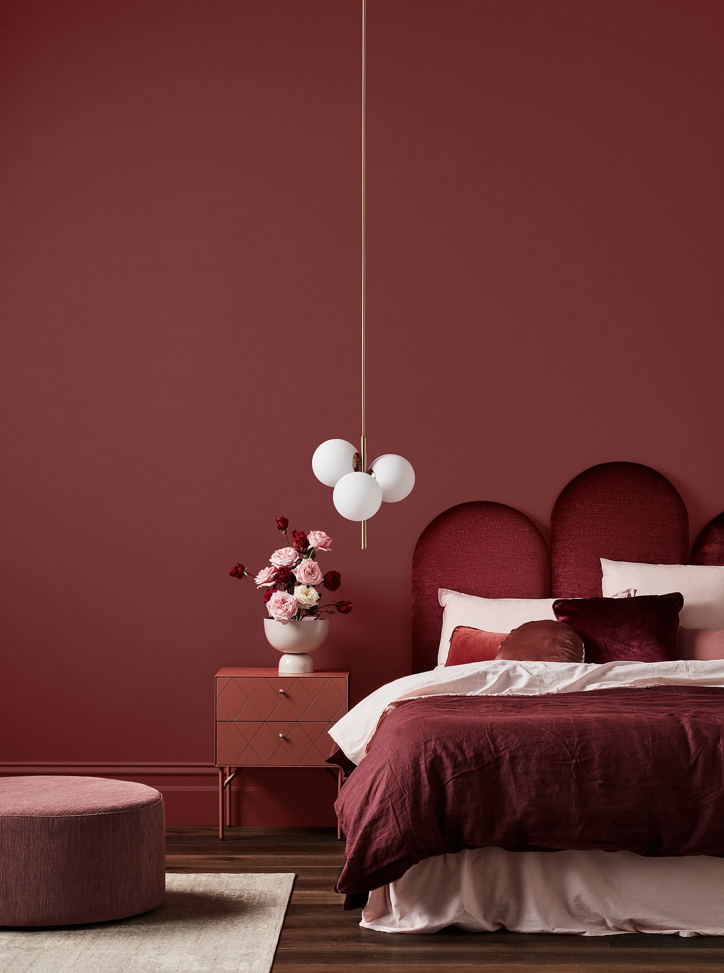 Deep red-pink bedroom with white pendant light, maroon velvet headboard, maroon and pink linens, timber bedside table and pink footrest.