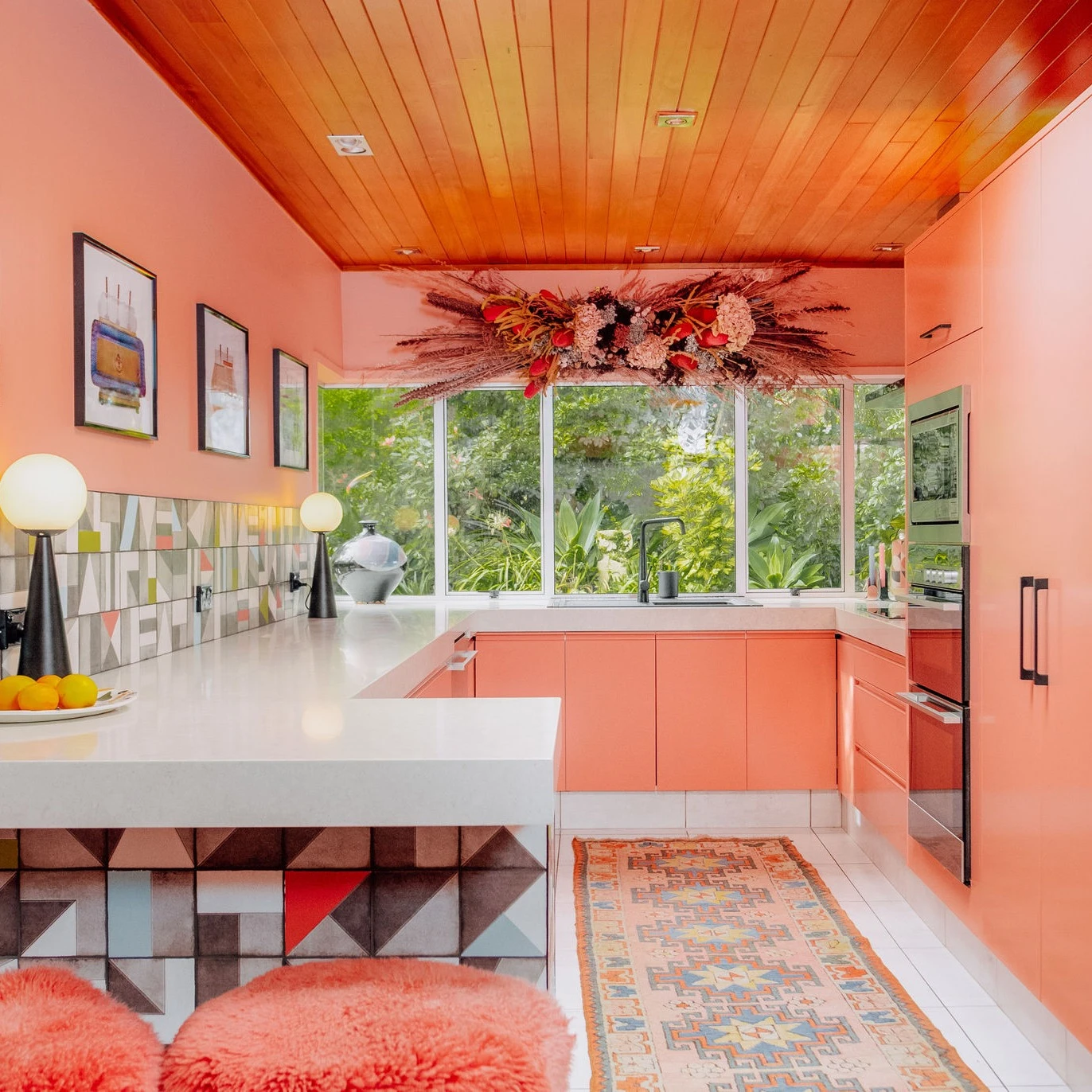 Evie Kemp's kitchen has pink walls and cabinets with a white benchtop. There is a large dried flower arrangement hanging above the benchtop and there are three pieces of art on the left wall.