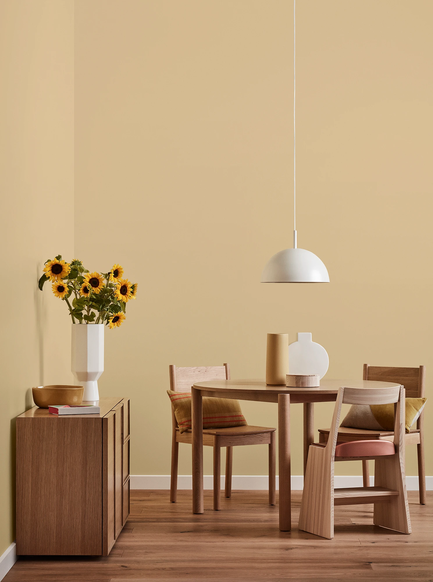 Round wooden table, three chairs and console with vase of sunflowers and white pendant light in yellow-brown room.