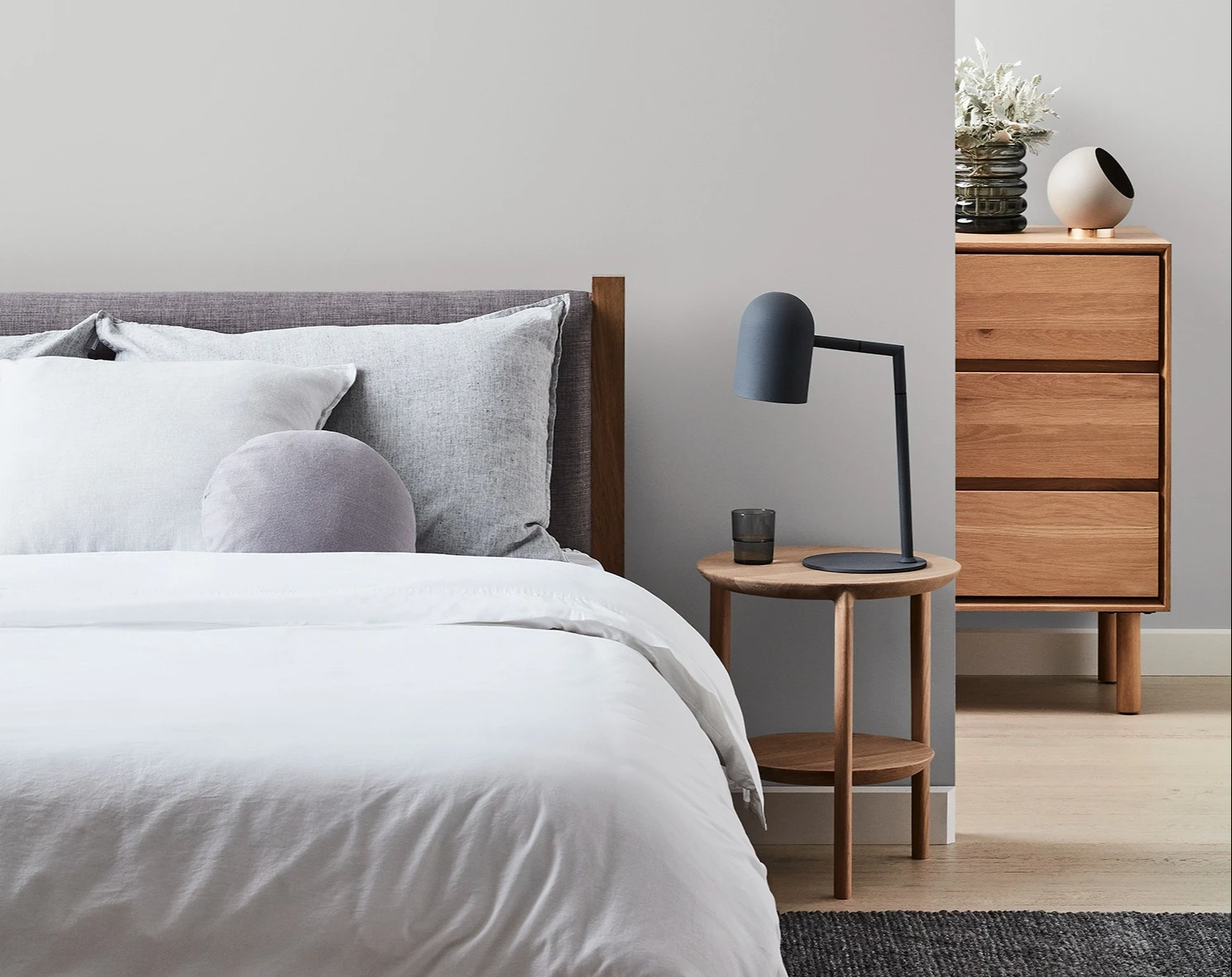 Grey painted wall with bed against it and side table with lamp. Tallboy showing partially behind wall.