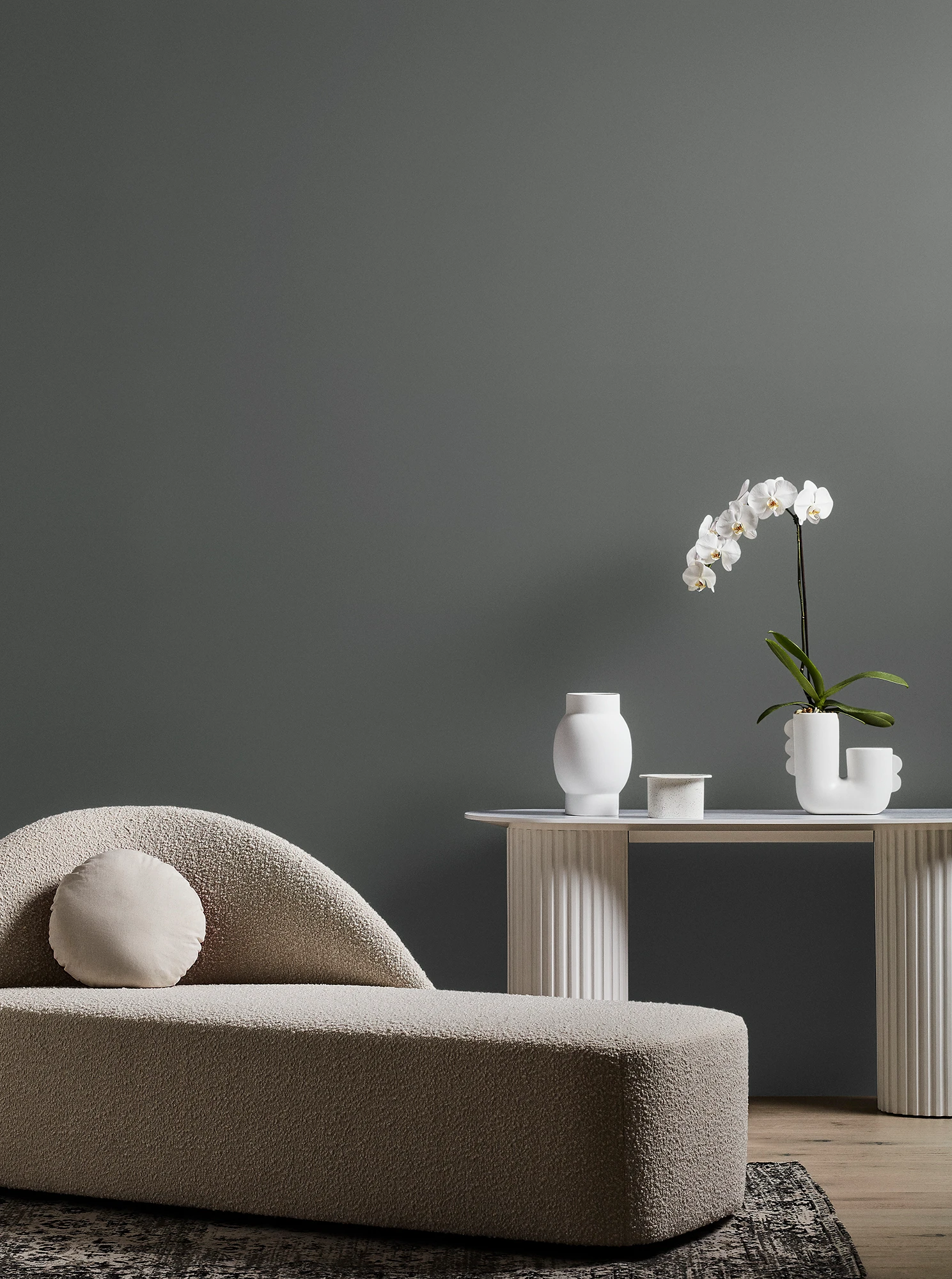  Neutral-coloured modern chaise longue and white table with orchid plant and vase in grey living room.