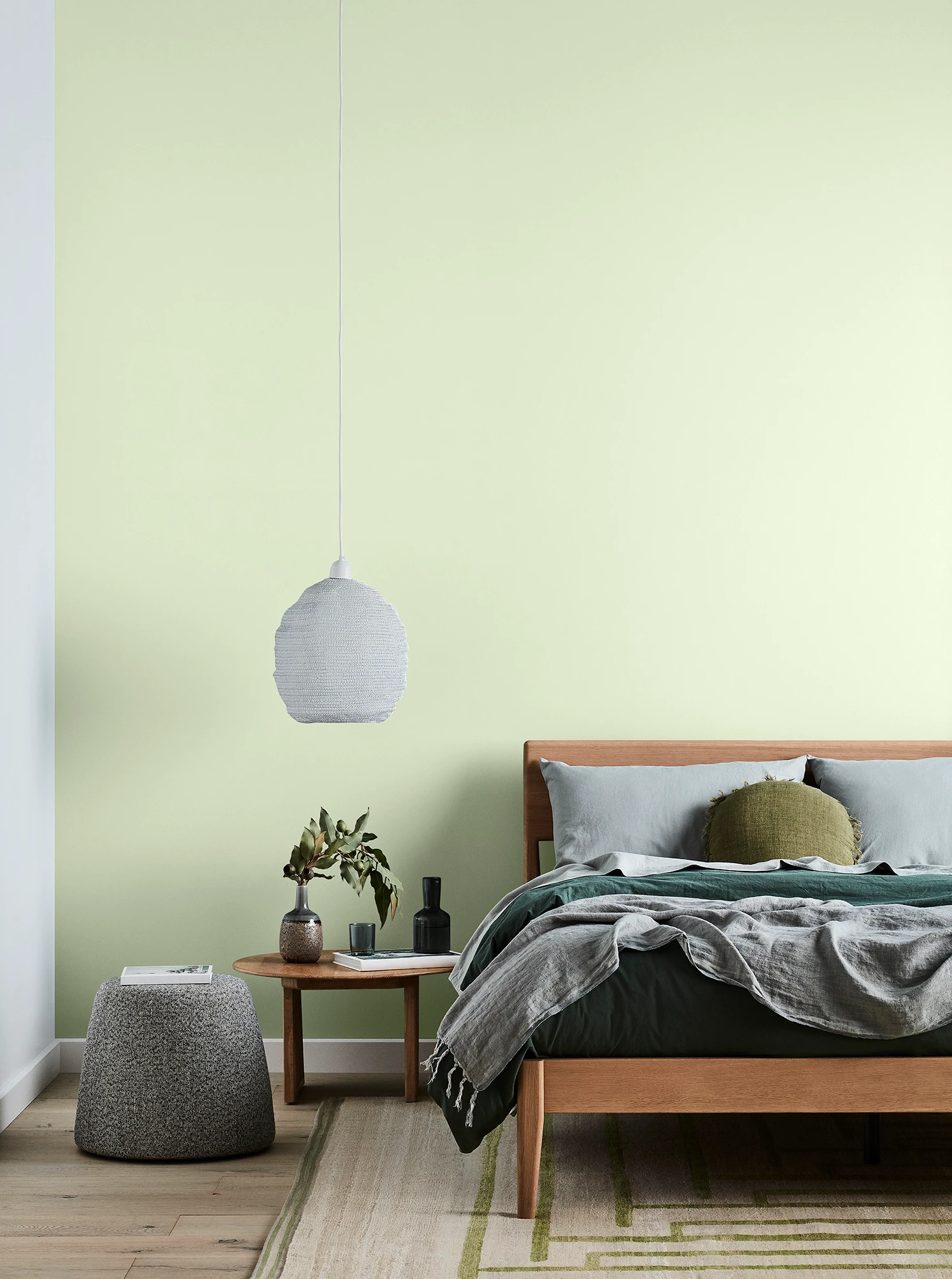 Green bedroom with timber bed and side table, white pendant light and grey linens.