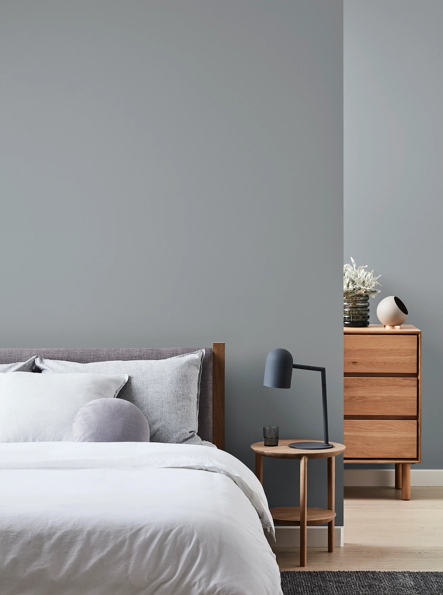 Grey bedroom with wooden chest of drawers and bedside table with black lamp and neutral coloured bed linen.