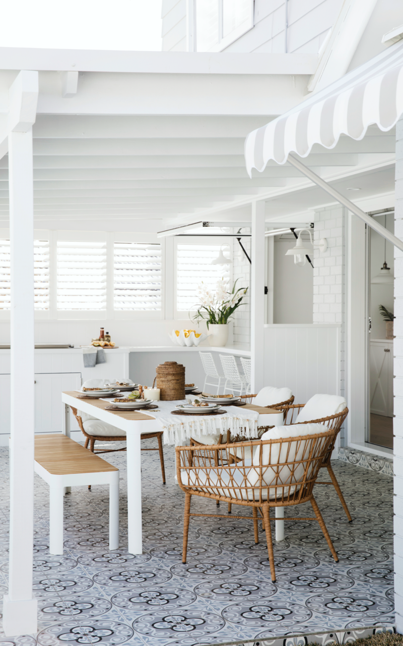 White alfresco area with white table, white bench seat and cane chairs. Colour Me Hamptons project by Three Birds Renovations.