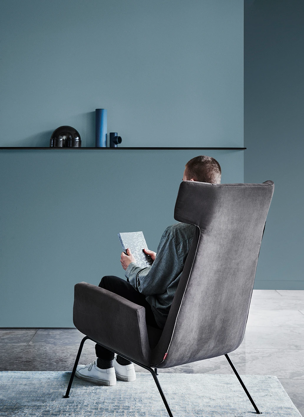 Man reading in high-backed modern armchair in room with blue-green walls and polished concrete.