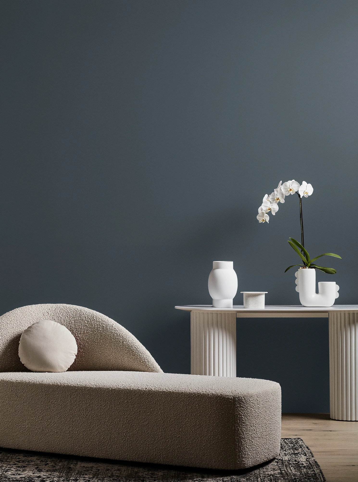 Neutral-coloured modern chaise longue and white table with orchid plant and vase in grey living room.