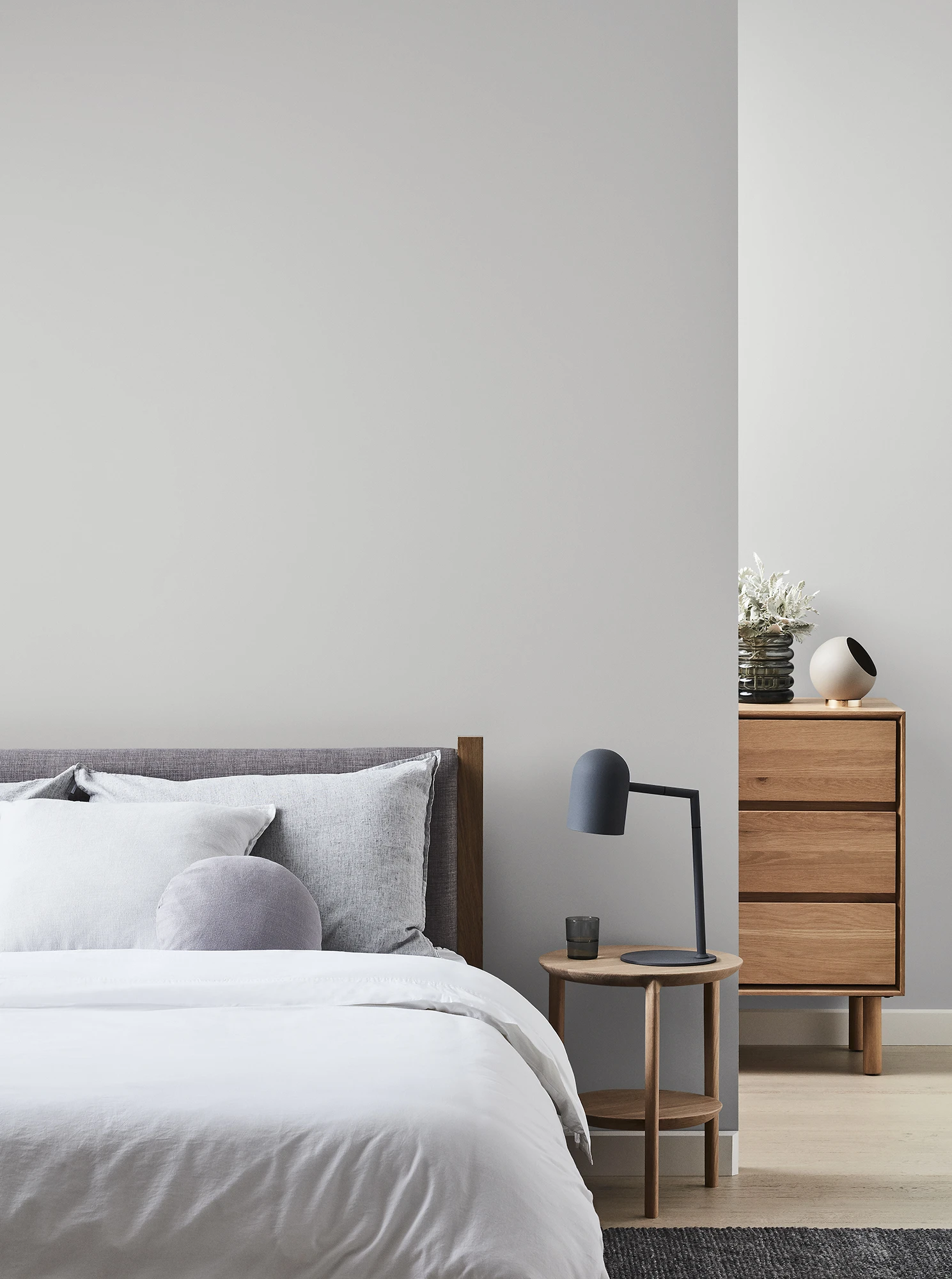 Grey painted wall with bed against it and side table with lamp. Tallboy showing partially behind wall.