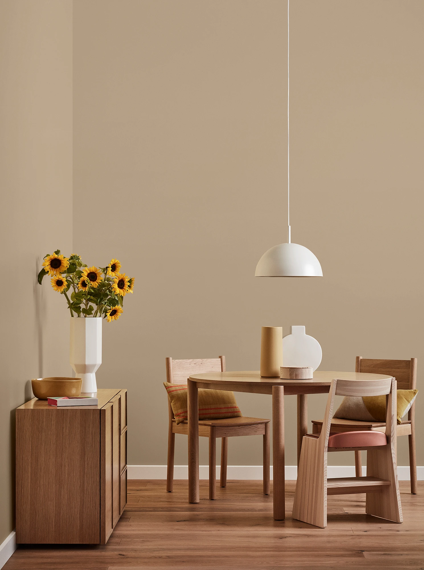 Round wooden table, three chairs and console with vase of sunflowers and white pendant light in neutral room.