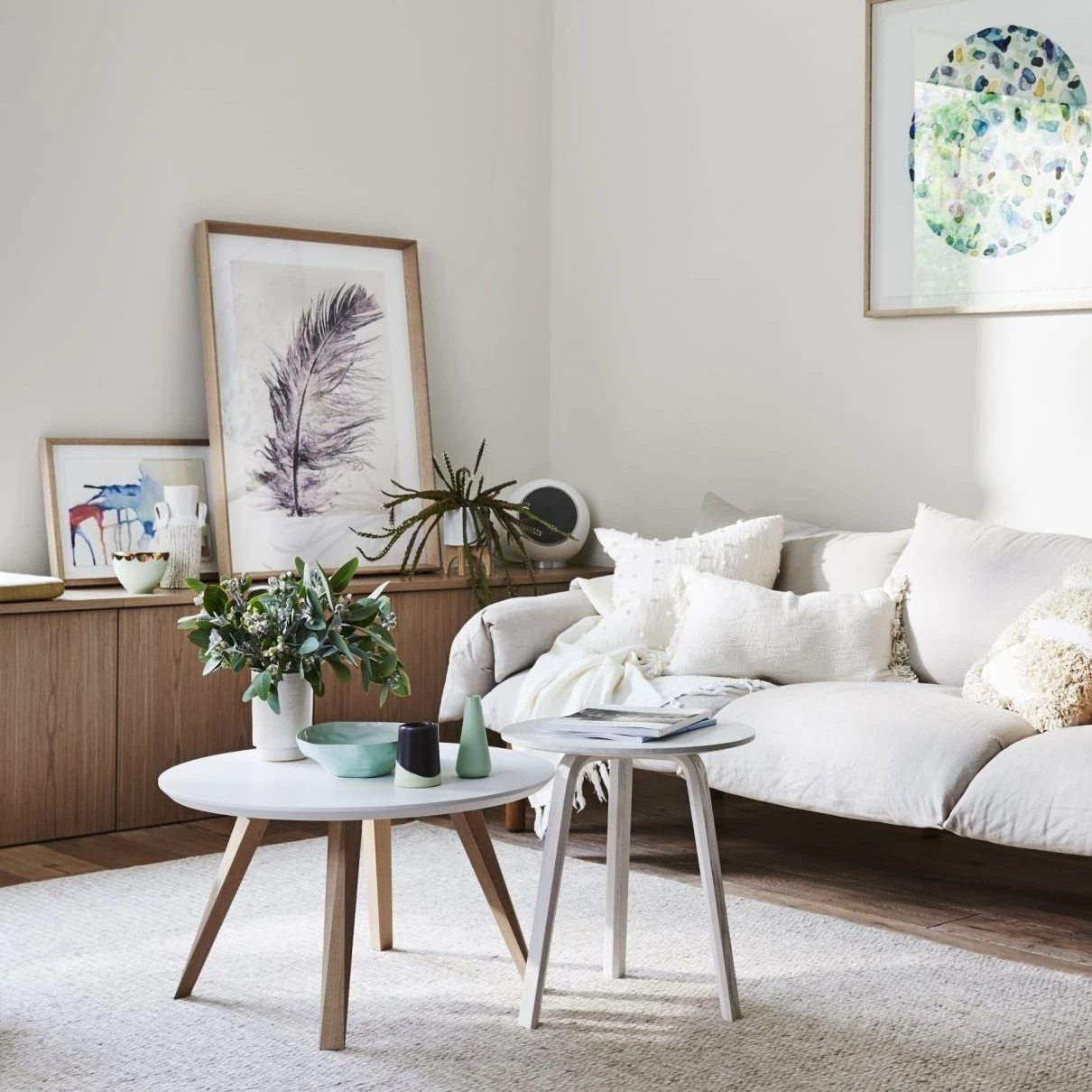 White couch and stacked coffee table in lounge room. Wooden buffet against background wall with paintings sitting on top and a flower painting on the wall. 