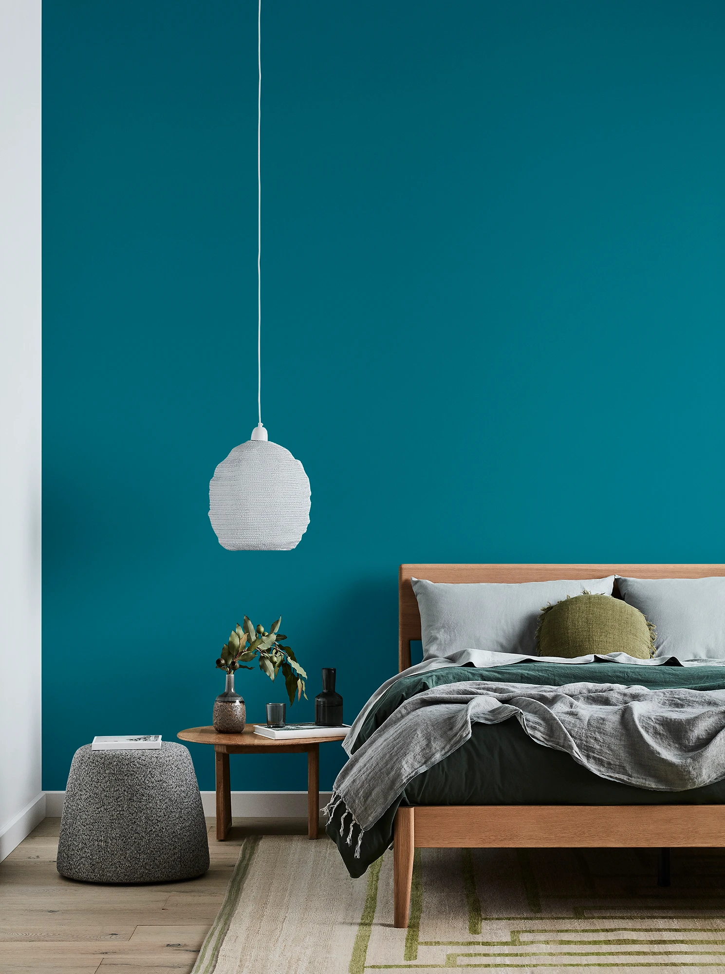 Green bedroom with timber bed and side table, white pendant light and grey linens.