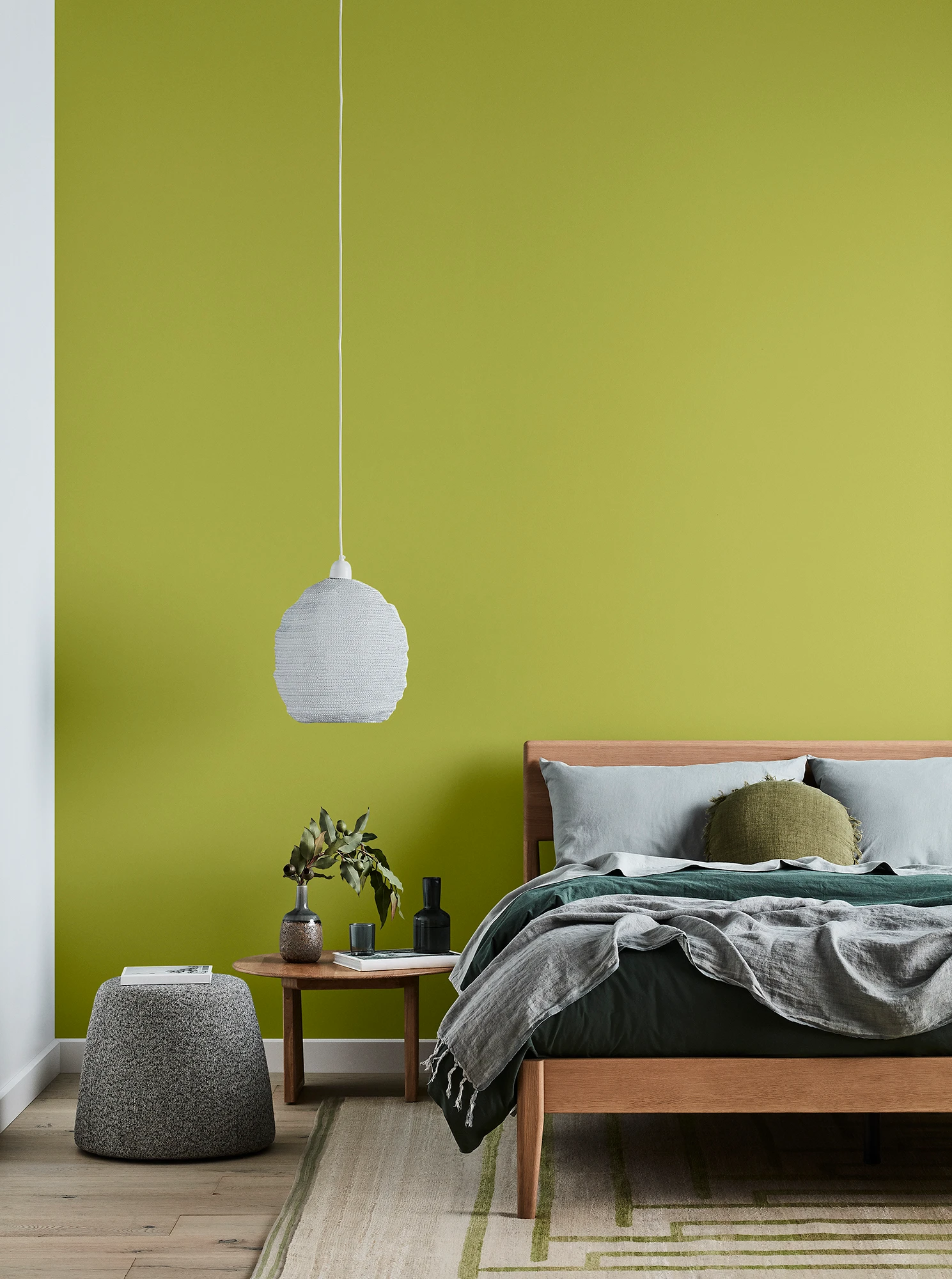 Green bedroom with timber bed and side table, white pendant light and grey linens.