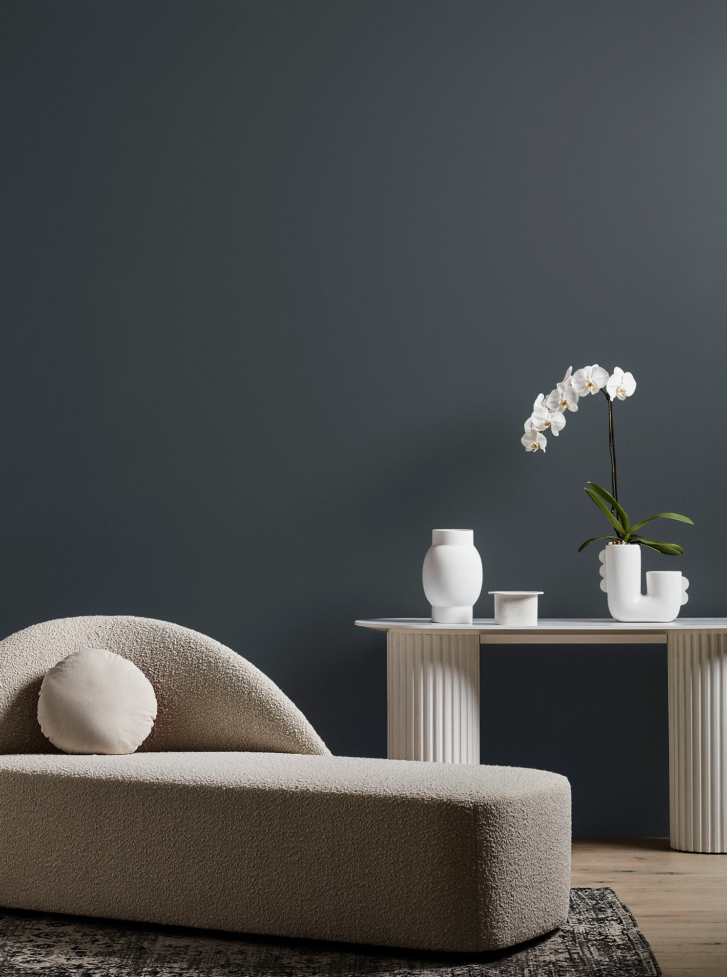 Neutral-coloured modern chaise longue and white table with orchid plant and vase in grey living room.