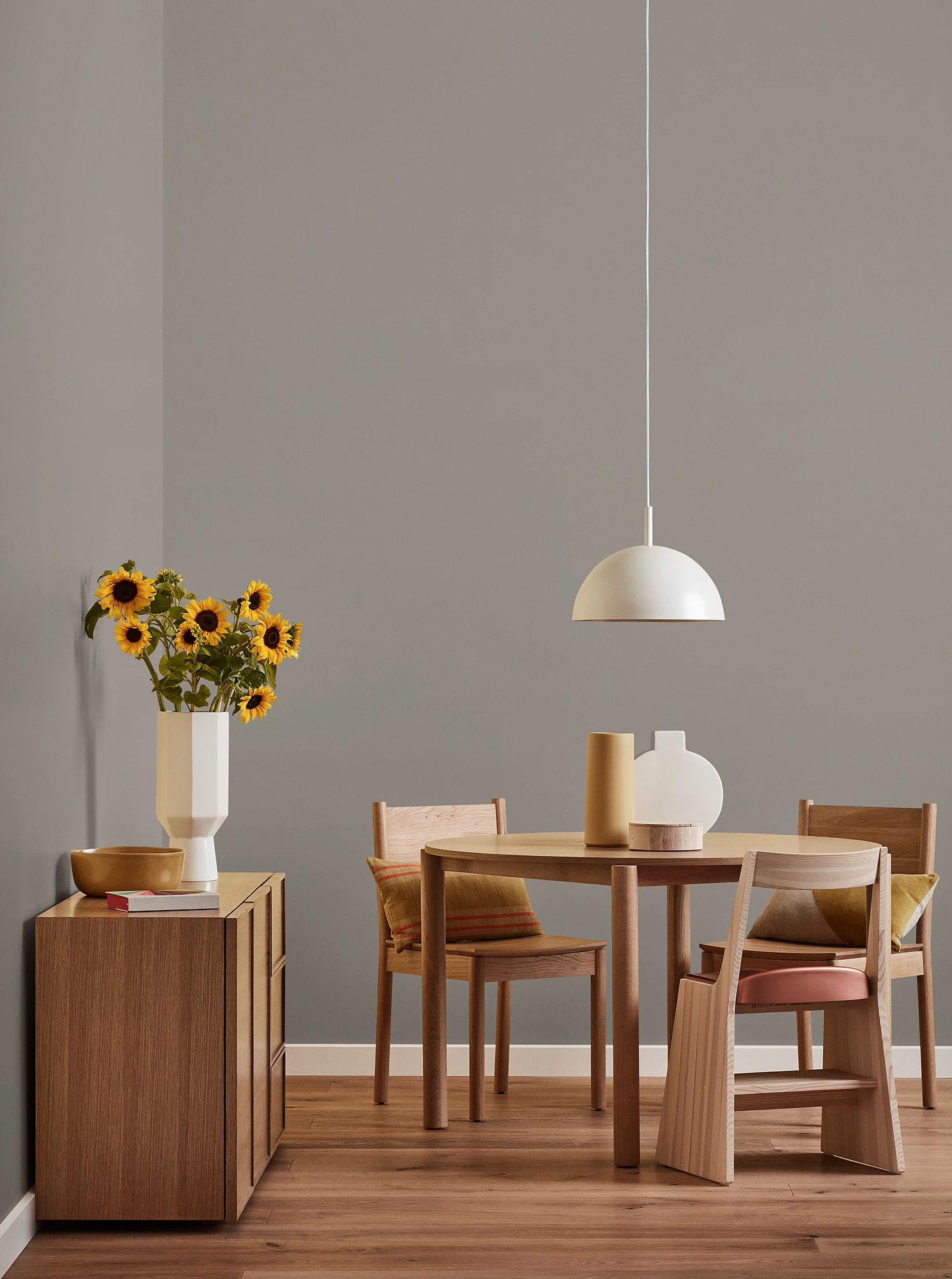 Round wooden table, three chairs and console with vase of sunflowers and white pendant light in neutral room.