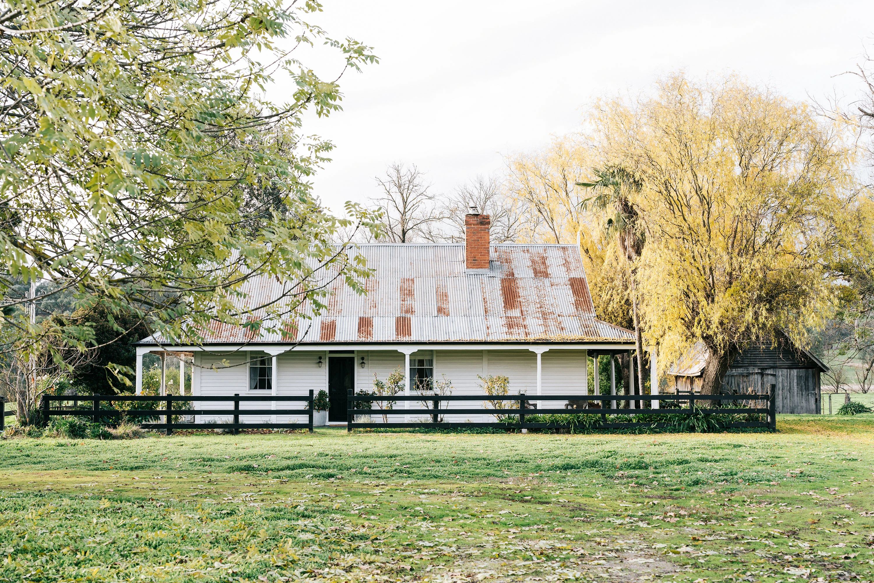 White farmhouse cottage with rusty corrugated roof and black fence