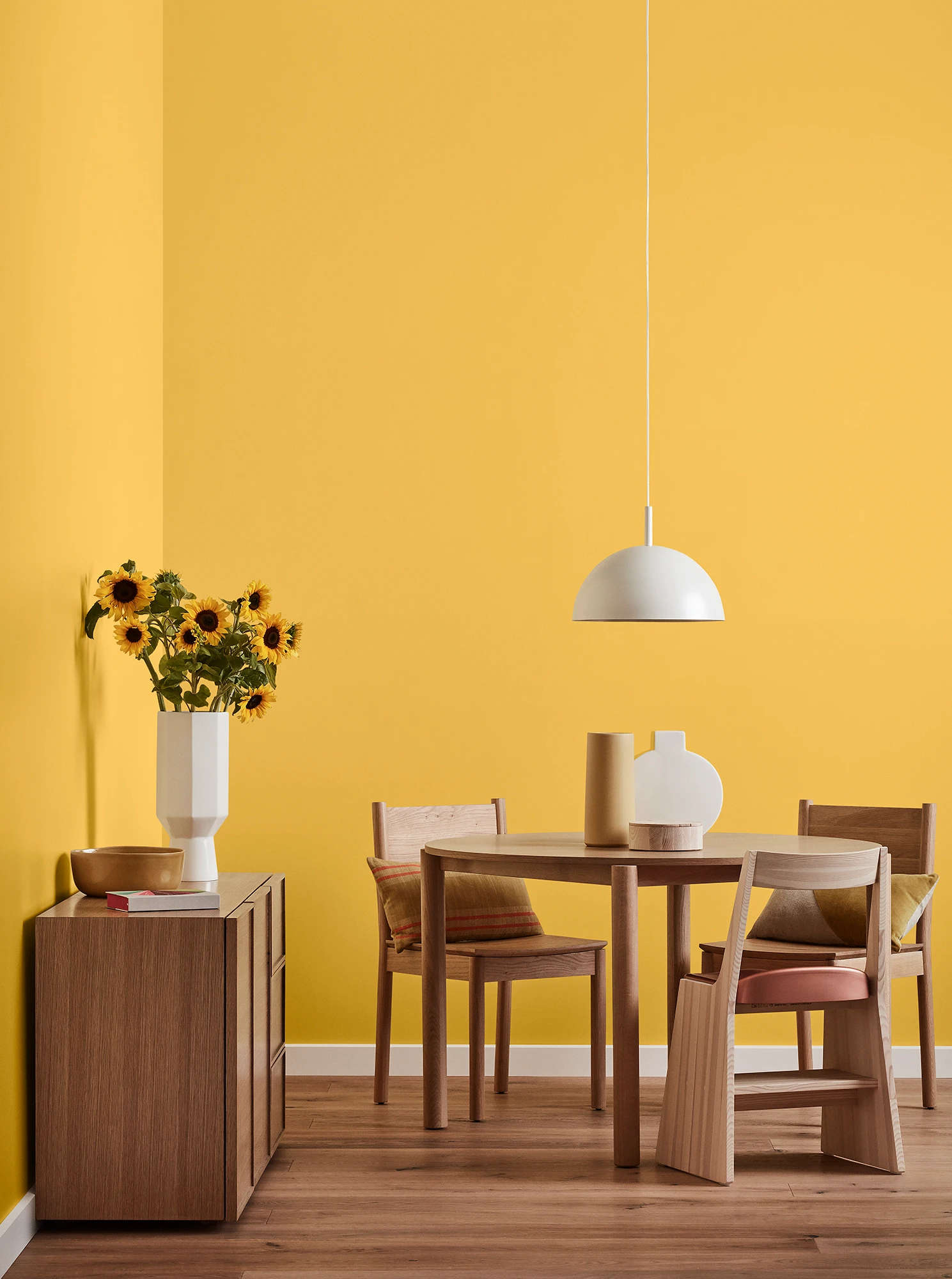 Round wooden table, three chairs and console with vase of sunflowers and white pendant light in a yellow room.