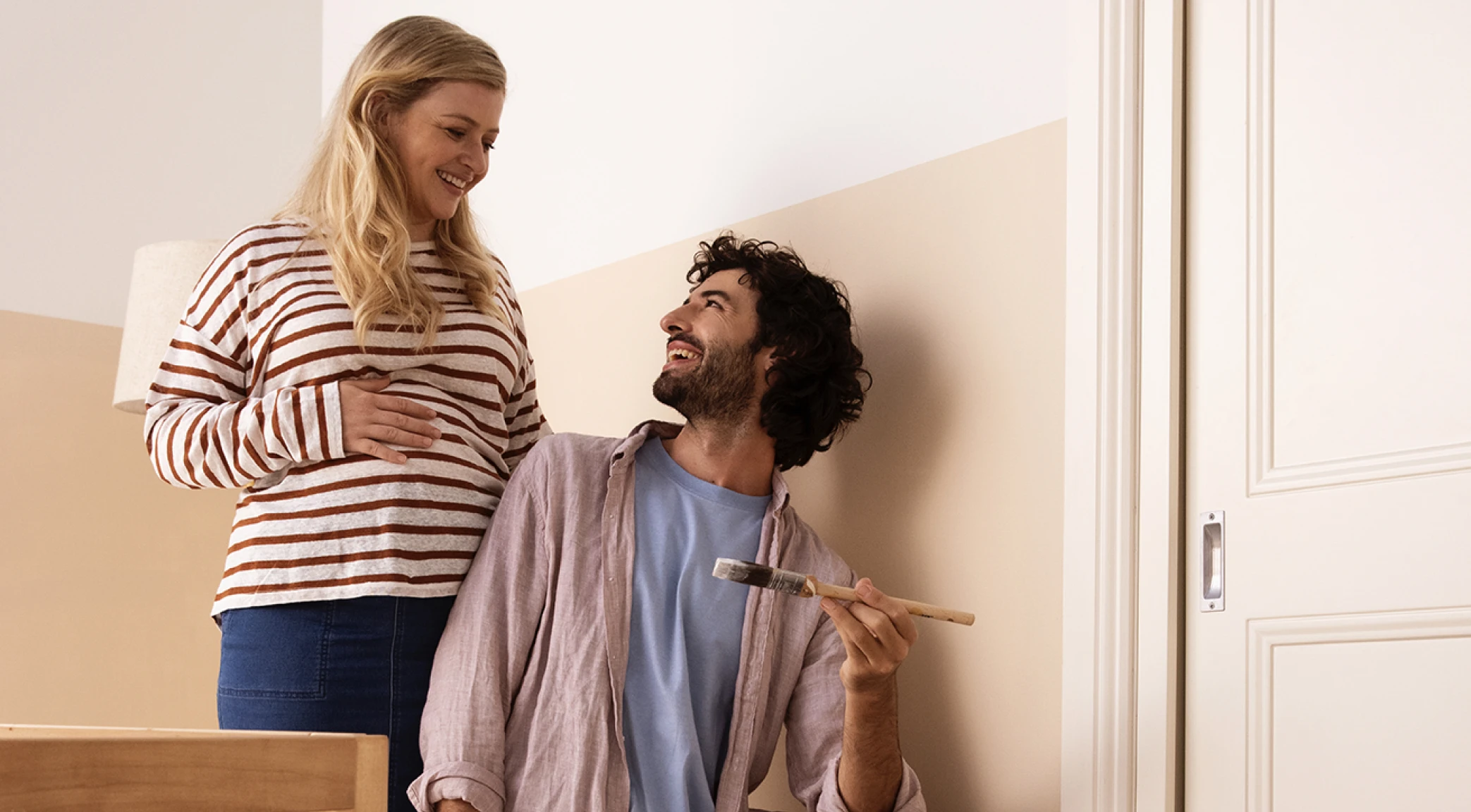 Husband painting a beige half wall in preparation for a baby with pregnant wife standing behind him.