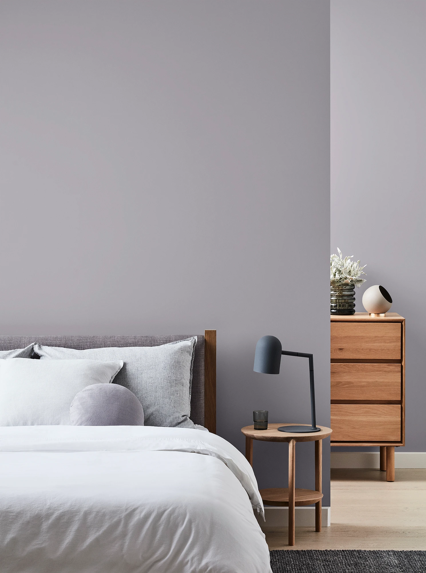 Grey bedroom with wooden chest of drawers and bedside table with black lamp and neutral coloured bed linen.