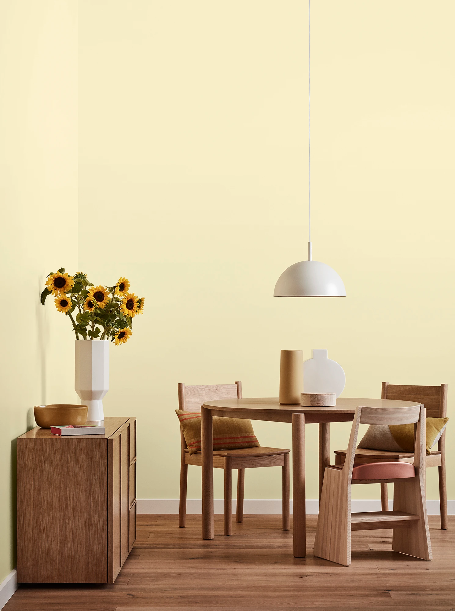 Round wooden table, three chairs and console with vase of sunflowers and white pendant light in yellow room.