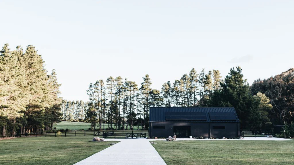 Distant view of black barn with trees and paddocks in background.