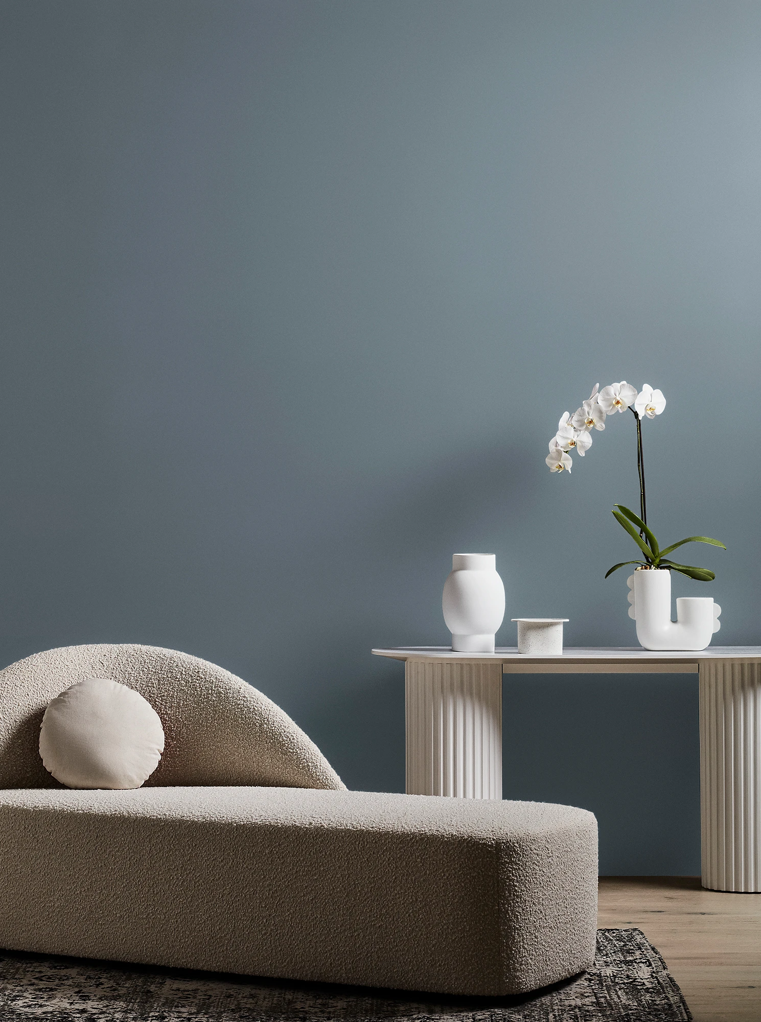 Neutral-coloured modern chaise longue and white table with orchid plant and vase in grey living room.