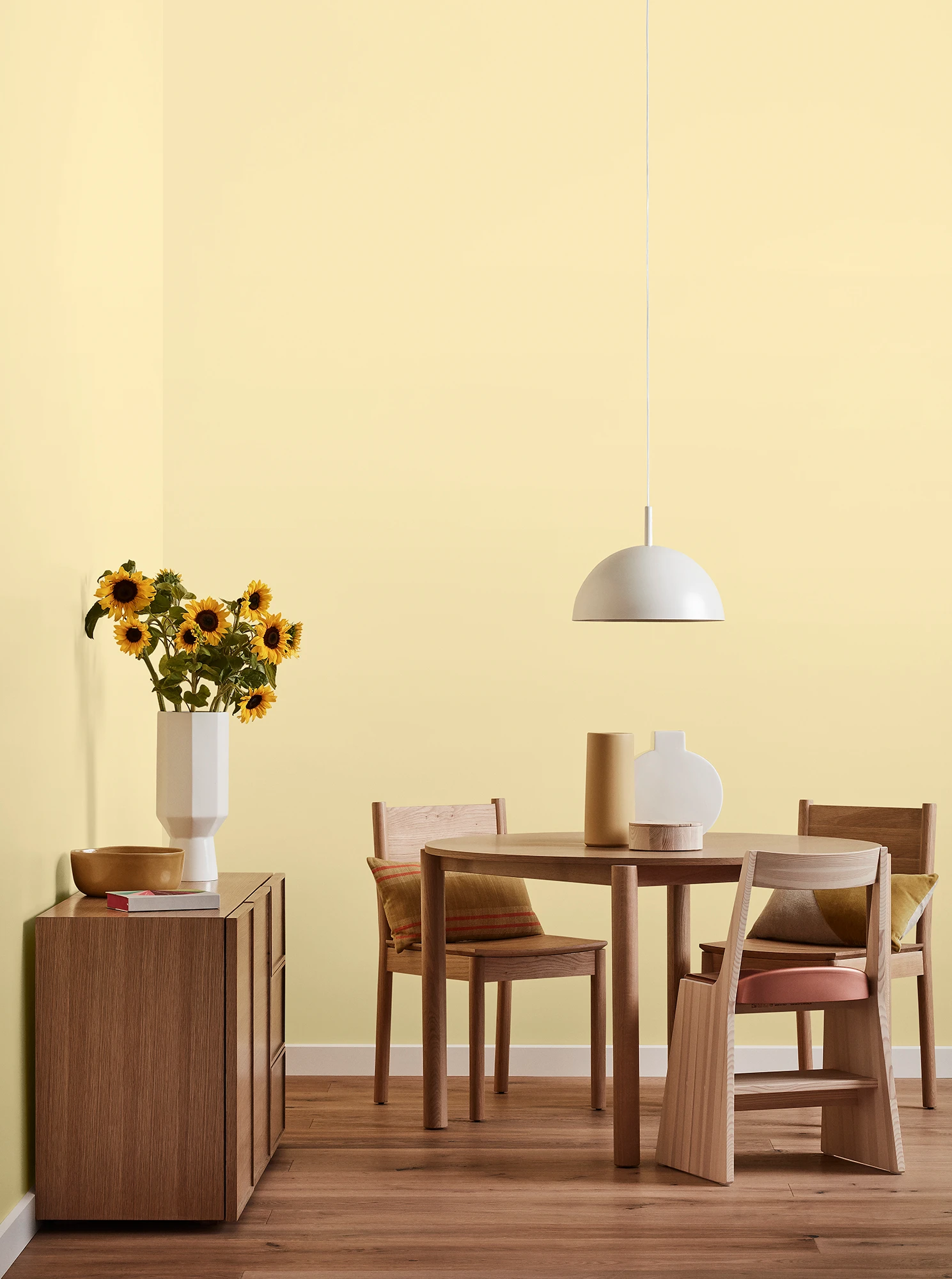  Round wooden table, three chairs and console with vase of sunflowers and white pendant light in light yellow oom.