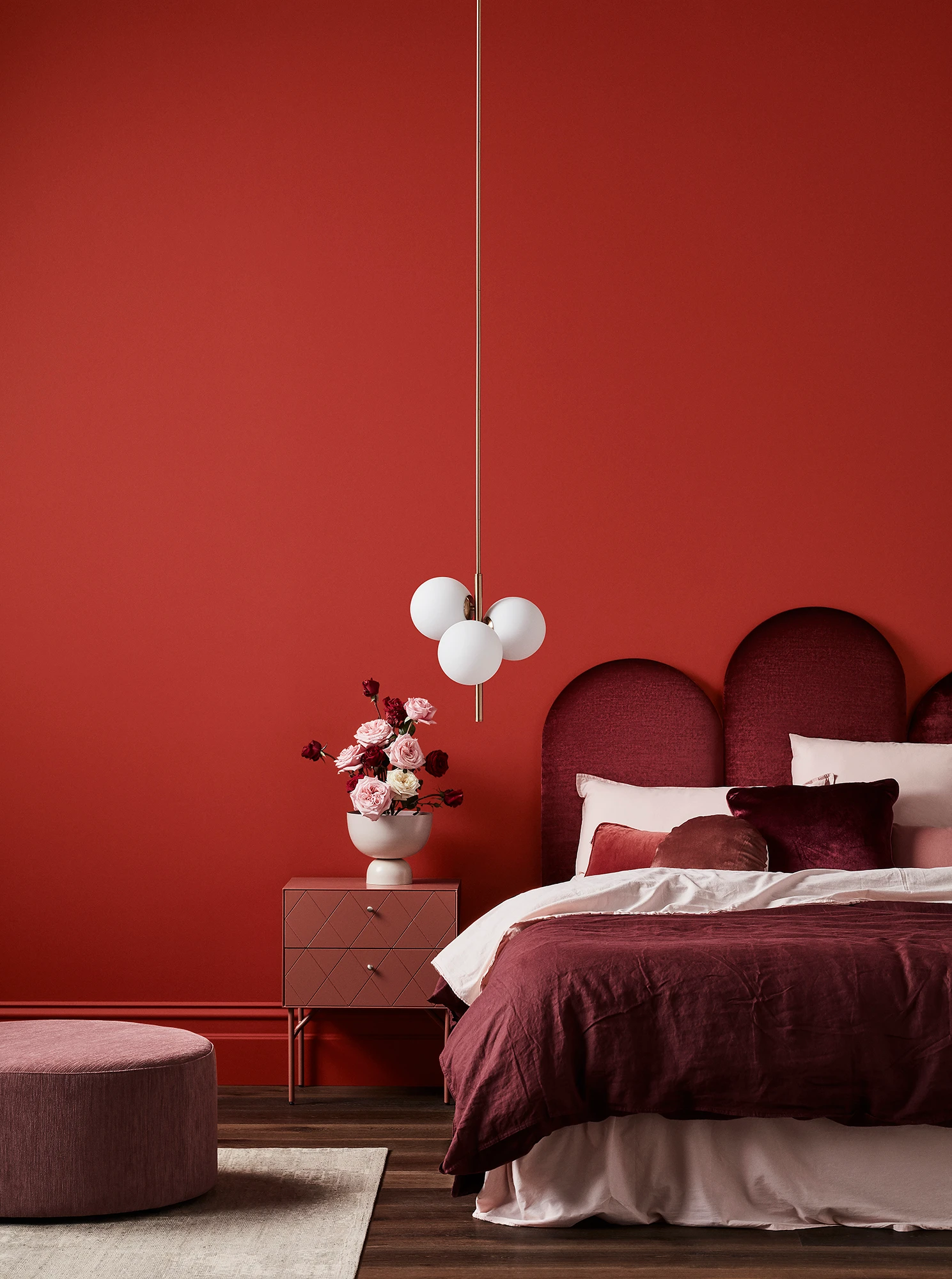 Red bedroom with white pendant light, maroon velvet headboard, maroon and pink linens, timber bedside table and pink footrest.