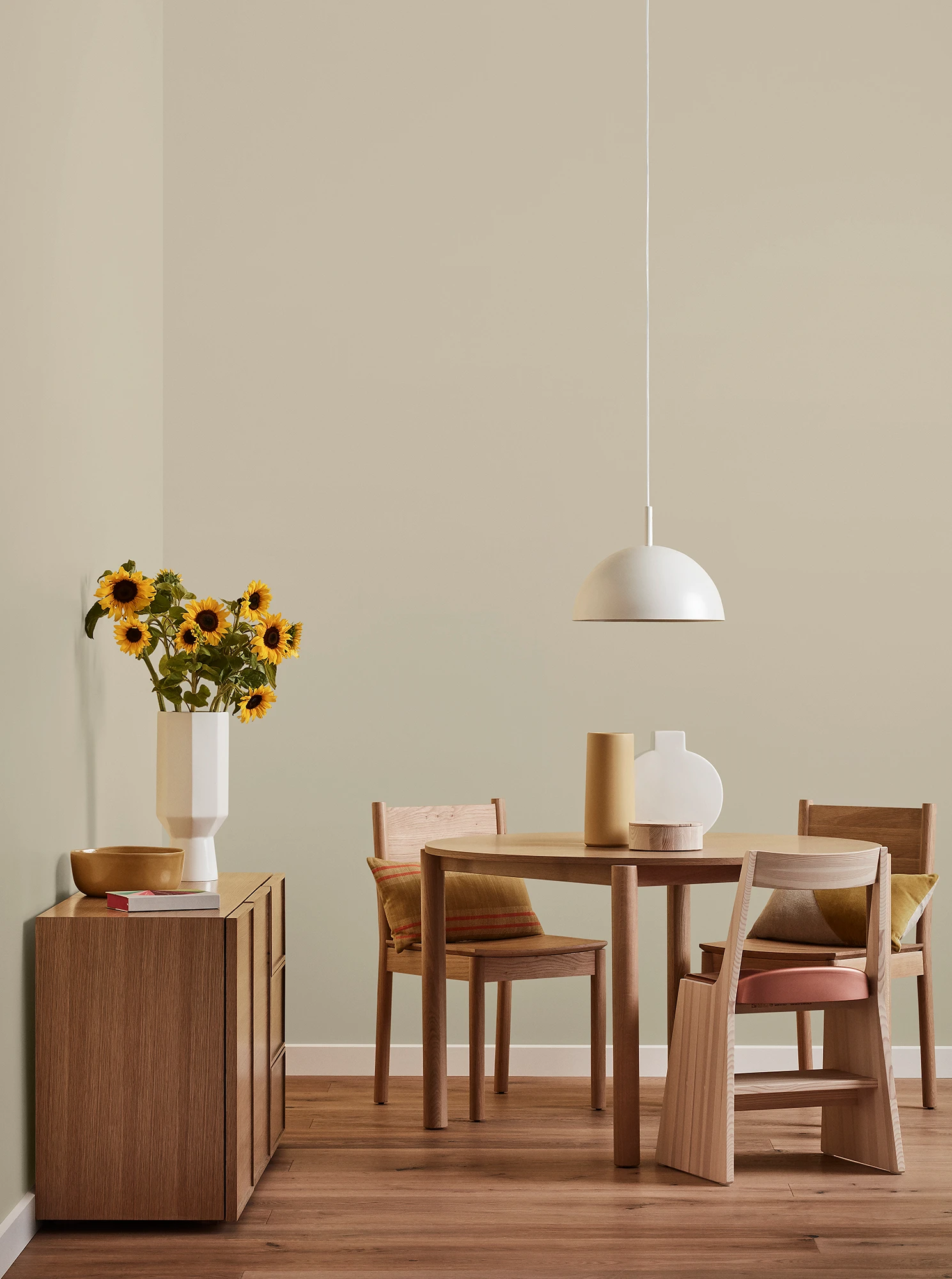 Round wooden table, three chairs and console with vase of sunflowers and white pendant light in neutral room.