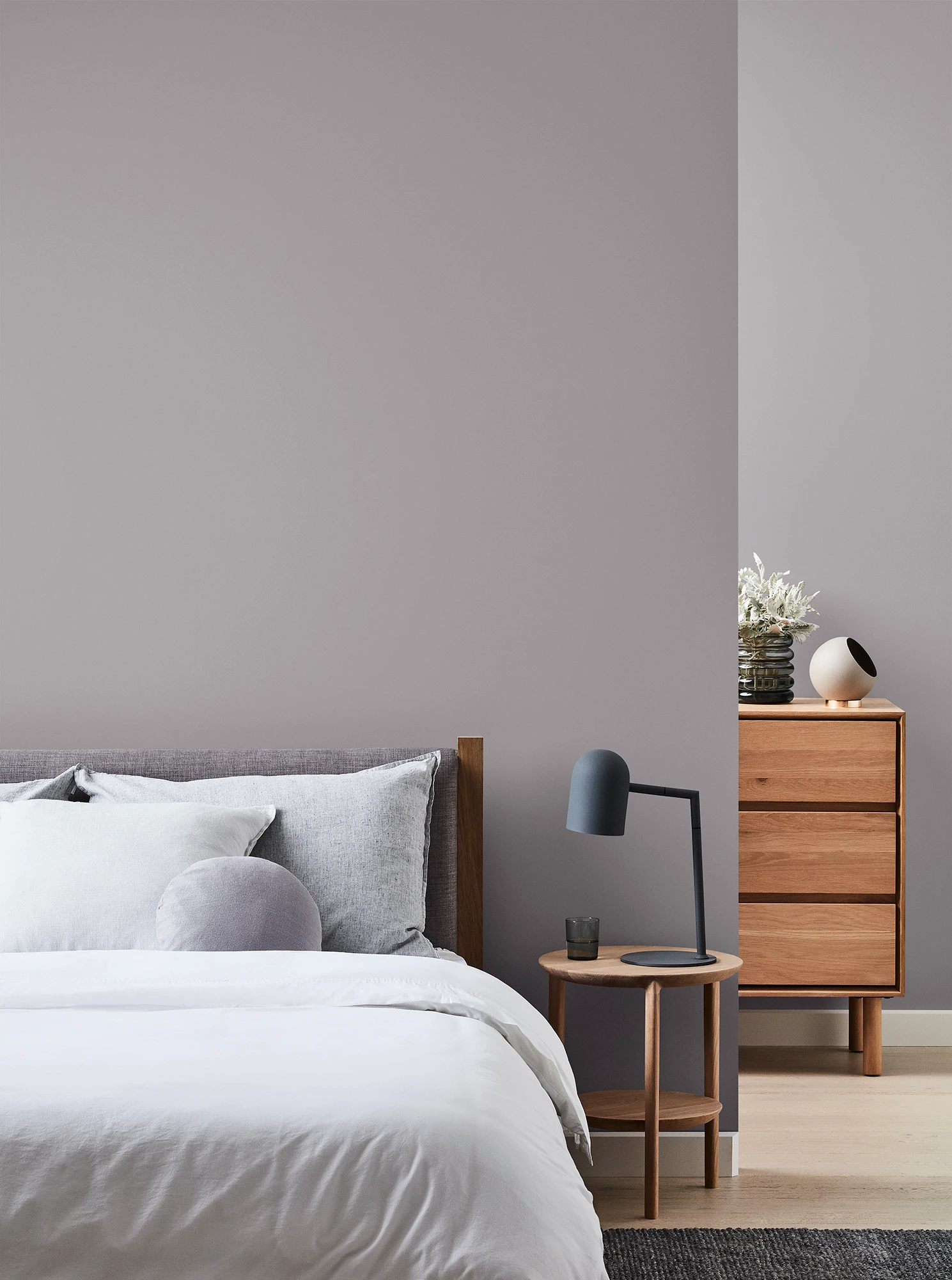 Grey bedroom with wooden chest of drawers and bedside table with black lamp and neutral coloured bed linen.